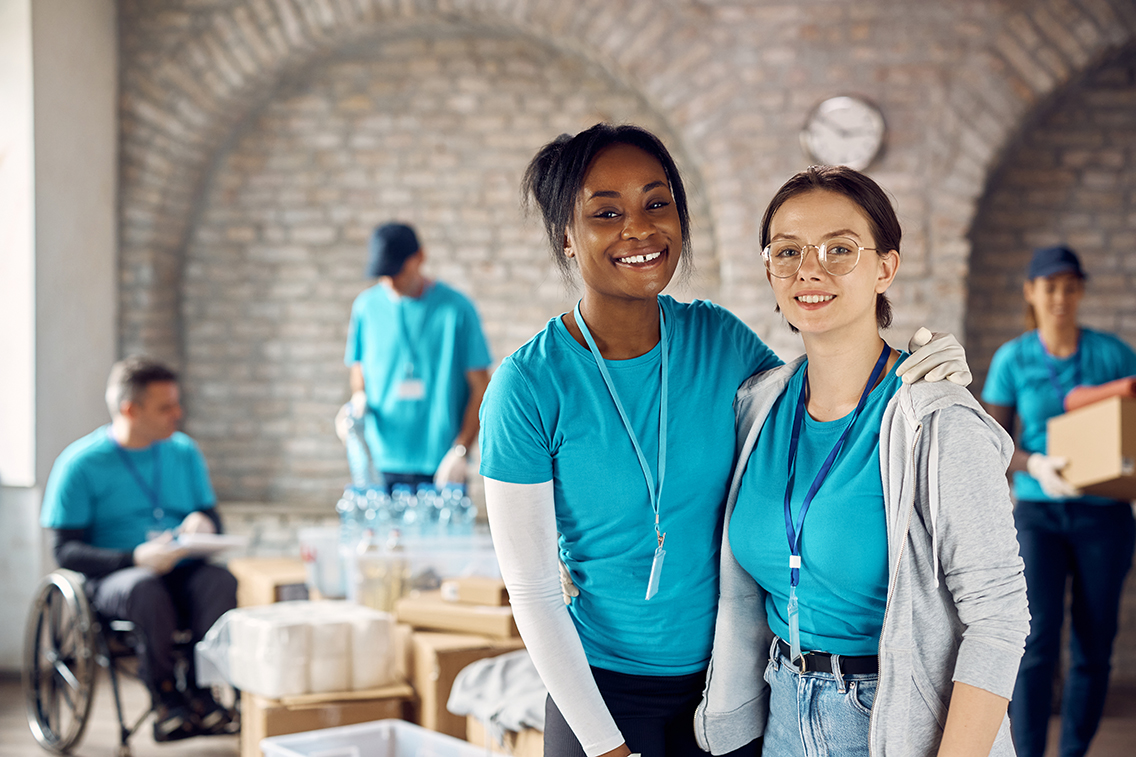 Two women working as volunteers at humanitarian aid center and smiling at camera.