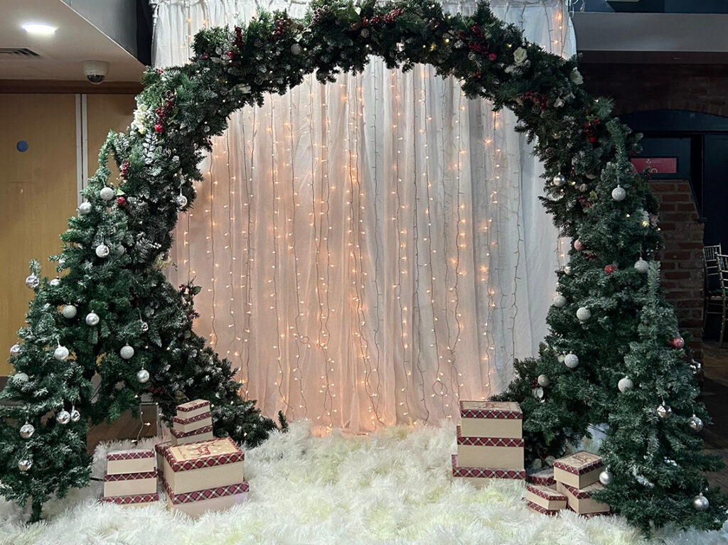 A designated photo area with a winter-themed backdrop of twinkling lights, a frosted wreath arch, gift boxes, and a faux snow carpet