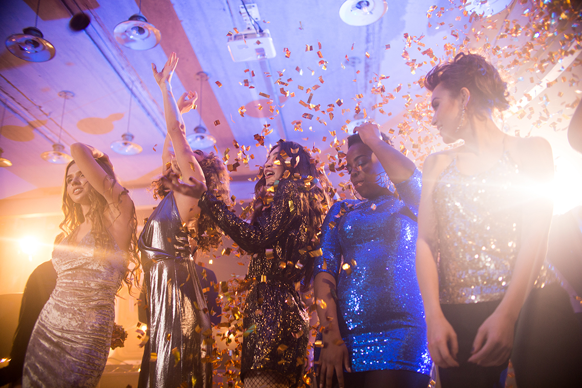 Group of beautiful young women wearing glittering dresses dancing under golden confetti enjoying party